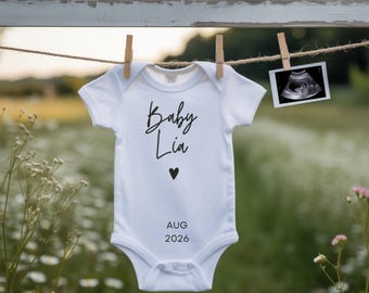 以下が含まれることがあります： 「Baby Lia」の文字が筆記体で書かれた白いベビー用ロンパース。黒いハートと「AUG 2026」の日付がプリントされています。 ロンパースは、超音波画像と一緒に洗濯ロープに吊るされています。