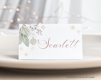 Puede incluir: Una tarjeta de lugar blanca con un diseño invernal que presenta hojas verdes, copos de nieve dorados y un adorno rojo y blanco. El nombre "Scarlett" está escrito en cursiva en un color rosa claro.