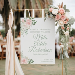 Könnte beinhalten: Ein weißes Willkommensschild mit floralen Akzenten und dem Text "WE ARE SO EXCITED TO OFFICIALLY WELCOME Mila Adele Robertson TO OUR FAMILY!". Das Schild ist mit rosa und weißen Rosen und Grün geschmückt. Das Datum "19 APRIL 2026" und der Ort "LOS ANGELES, CA" sind ebenfalls angegeben.