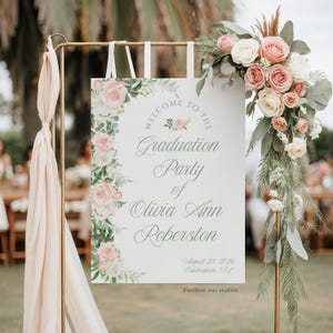 Pode incluir: Uma placa branca de festa de formatura com detalhes florais e o texto "Welcome to the Graduation Party of Olivia Ann Roberston, August 23, 2026, Celebration, FL." A placa é adornada com rosas rosa e brancas e vegetação. A placa é suportada por um suporte de metal dourado.