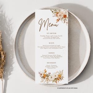 Könnte beinhalten: Ein herbstliches Menü mit einem floralen Design mit Kürbissen, Blättern und Beeren. Das Menü listet Vorspeisen, Hauptgerichte, Beilagen und Desserts auf. Der Text lautet: "Menü, Vorspeise, Kürbiscremesuppe mit Crème fraîche, Prosciutto-umwickelte Feigen, Verschiedene Brote und Käse, Hauptgericht, Kräuter-gekröntes Lammkarree, Gebratener Lachs mit Zitronenbutter, Filet Mignon mit Sauce Hollandaise, Beilagen, Gebratene Kartoffeln mit Thymian, Süßkartoffeln mit Marshmallows, Grüne Bohnen mit Schalotten, Dessert, Klassischer Tiramisu, Kürbis-Käsekuchen, Verschiedene Torten, FARTHEST STAR STUDIOS."