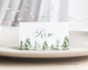 Puede incluir: Una tarjeta de sitio blanca con el nombre "Rose" en escritura elegante. El borde inferior presenta un diseño de acuarela de árboles de hoja perenne verdes y blancos. La tarjeta está colocada sobre un plato color crema, con un fondo borroso.