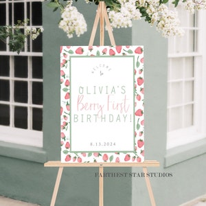 May include: Welcome to Olivia's Berry First Birthday! A white sign with a green border and a strawberry pattern. The text is in pink and green. The date 8.13.2024 is printed below the text. The sign is on a wooden easel.