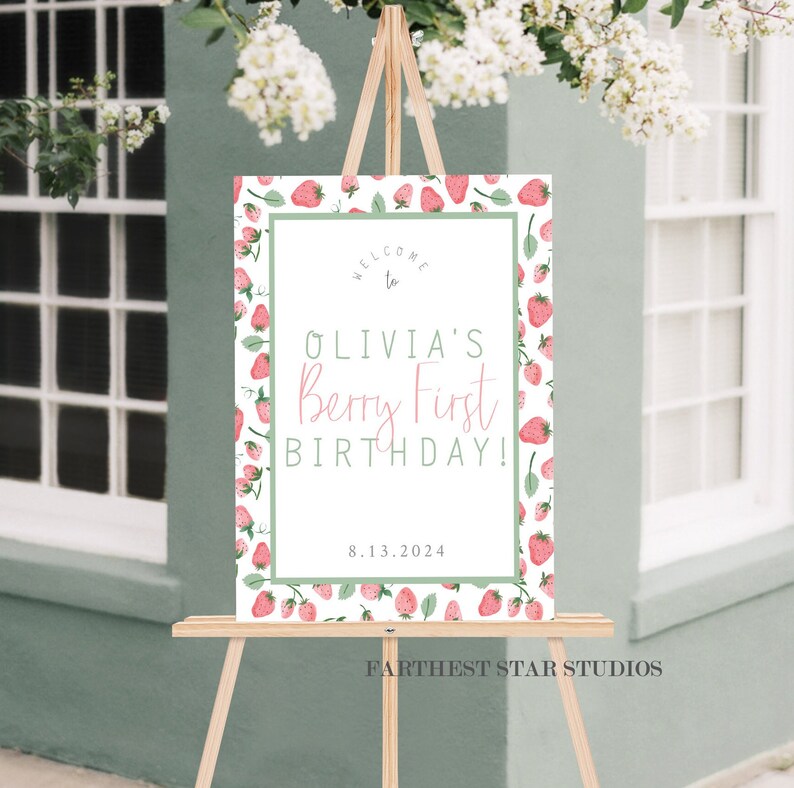 May include: Welcome to Olivia's Berry First Birthday! A white sign with a green border and a strawberry pattern. The text is in pink and green. The date 8.13.2024 is printed below the text. The sign is on a wooden easel.