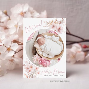 Könnte beinhalten: Eine rosa-weiße florale Geburtsanzeige mit einem Foto eines schlafenden Babys in einem Korb. Die Karte sagt "Willkommen!" und "Adele Marie" mit dem Geburtsdatum, dem Gewicht und der Größe des Babys. Die Karte ist von "Farthest Star Studios."