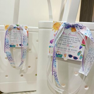 May include: Two white tote bags with handles, each with a colorful summer-themed gift tag attached. The tags have a cheerful message thanking the recipient for their hard work and wishing them a happy summer. The tags are tied with a purple and white ribbon.