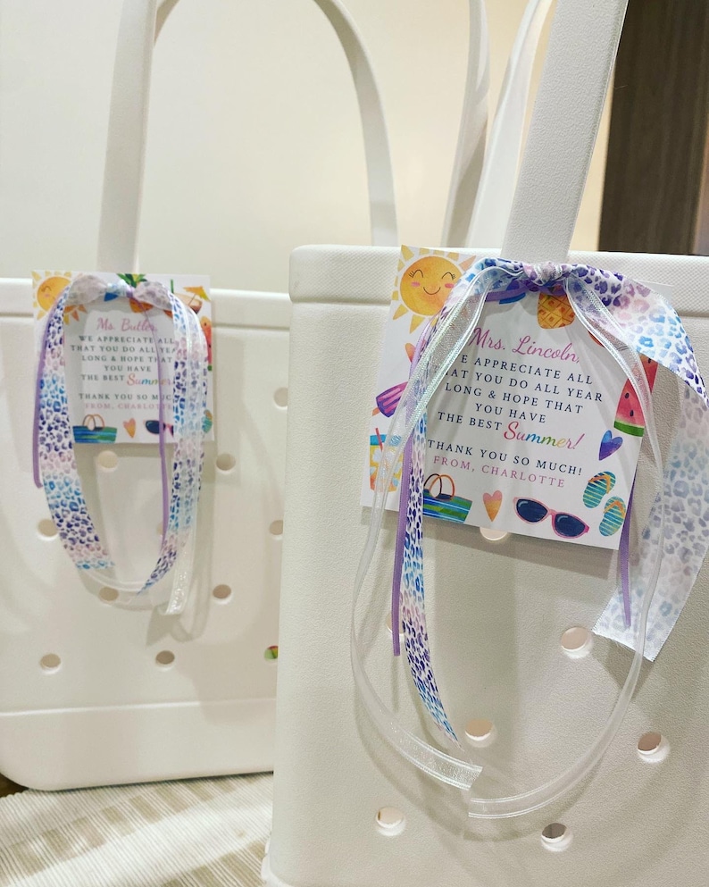 May include: Two white tote bags with handles, each with a colorful summer-themed gift tag attached. The tags have a cheerful message thanking the recipient for their hard work and wishing them a happy summer. The tags are tied with a purple and white ribbon.