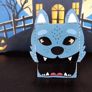 May include: A blue wolf-faced paper box with a black mouth and white teeth. The wolf has white-tipped ears and yellow eyes. The background features a Halloween scene with a spooky house, fence, and jack-o'-lanterns.