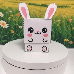Può includere: Un coniglietto di carta bianco con l'interno delle orecchie, le guance e i cuscinetti delle zampe rosa. Il coniglietto ha grandi occhi neri e un piccolo naso e bocca neri. Il coniglietto è su una superficie bianca con uno sfondo sfocato di un campo di fiori.