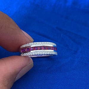 Peut inclure: Une bague en argent ornée d'une rangée centrale de pierres rouges vives taillées en carré, encadrée de deux rangées de petites pierres transparentes. La bague est présentée sur un fond bleu, soulignant son design détaillé et son éclat.