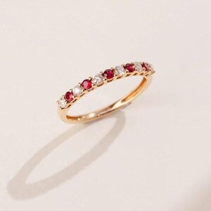 May include: A gold band ring featuring alternating red and clear gemstones. The ring is set with small, round gemstones, creating a continuous pattern around the band. The ring is displayed on a light-colored surface, with its shadow visible.
