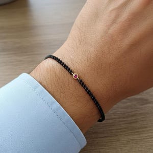Puede incluir: Una pulsera de cadena negra con una pequeña gema roja redonda engastada en un bisel dorado. La pulsera se lleva en la muñeca, con una manga de camisa azul claro visible.