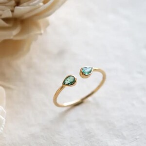 May include: Open-style gold ring with two teardrop-shaped, light green gemstones. The ring has a delicate, minimalist design. The gemstones are set in a gold band, suitable for everyday wear.