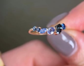 Puede incluir: Un anillo de oro con un grupo de piedras preciosas azules de varias formas y tonos. Las piedras preciosas incluyen cortes ovalados y redondos, creando un diseño único. El anillo es sostenido por una persona.