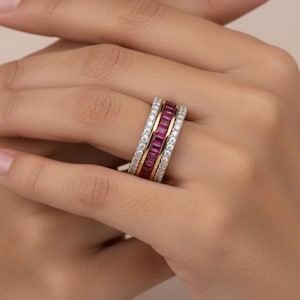 Peut inclure: Une bague à trois rangs avec un design or et argent. La bande centrale présente une rangée de pierres précieuses rectangulaires, rouge foncé. Les deux autres rangs sont bordés de petites pierres précieuses claires. La bague est sur le doigt d'une personne.