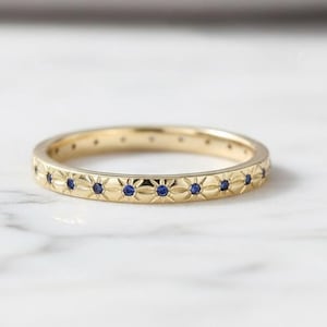 May include: A gold band ring with a repeating pattern of small blue gemstones. The ring has a floral design engraved around the band. The ring is displayed on a white marble surface.