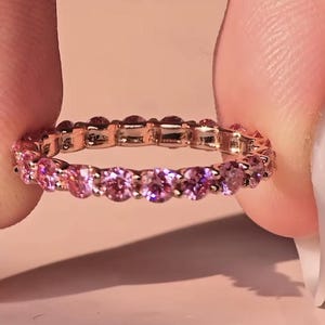Op de afbeelding: Een roségouden eternity ring met een doorlopende rij roze edelstenen. De ring wordt tussen twee vingers gehouden tegen een zachtroze achtergrond. De edelstenen zijn rond geslepen en variëren in roze tinten.