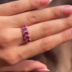 Könnte beinhalten: Ein goldfarbener Ring mit einer Reihe ovaler, leuchtend roter Edelsteine. Die Edelsteine sind in einer Reihe angeordnet, mit kleinen, klaren Steinen zwischen ihnen. Der Ring wird an einem Finger getragen und zeigt sein Design und seine Farbe.