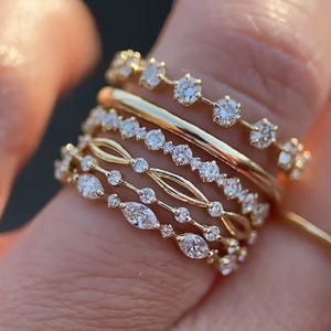 Puede incluir: Una colección de anillos de oro con varios diseños, incluyendo bandas con pequeñas piedras preciosas claras redondas y en forma de marquesa. Los anillos están apilados en un dedo, mostrando sus diferentes estilos y texturas. Los anillos son de oro.