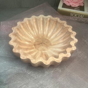 以下が含まれることがあります： スカラップエッジの装飾的な木製ボウル。ボウルは、大理石模様のライトブラウン色です。ピンクの花の形をしたキャンドルが背景の本の上にあります。