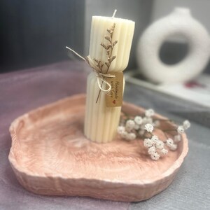 May include: A cream-colored, ribbed pillar candle with a dried floral accent and a small tag that reads "Handmade with love." The candle sits on a pink, organic-shaped tray, with a sprig of white flowers.