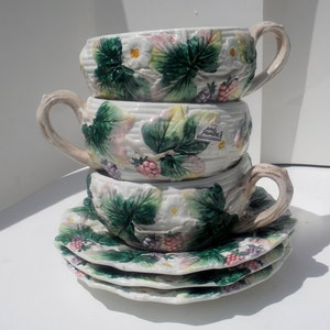 May include: A set of three vintage ceramic soup bowls with a hand-painted design of green leaves, white flowers, and pink and purple berries. The bowls are stacked on top of each other and have a label that reads "Hand Painted".