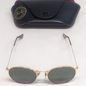 May include: Gold-framed sunglasses with oval lenses and a black case. The sunglasses have a classic design with a thin gold frame and dark green lenses. The case is black with a red interior and a logo.