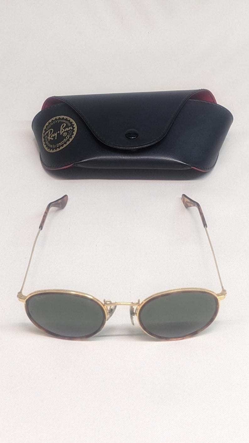 May include: Gold-framed sunglasses with oval lenses and a black case. The sunglasses have a classic design with a thin gold frame and dark green lenses. The case is black with a red interior and a logo.