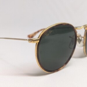 May include: Round sunglasses with a gold frame and dark green lenses. The frames have a tortoiseshell pattern on the outer edges. The temples are thin and silver, with a curved end. The sunglasses are set against a white background.