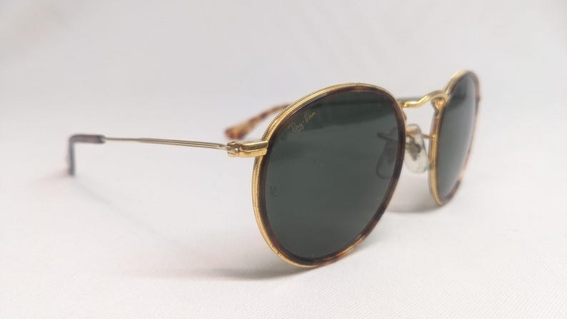 May include: Round sunglasses with a gold frame and dark green lenses. The frames have a tortoiseshell pattern on the outer edges. The temples are thin and silver, with a curved end. The sunglasses are set against a white background.