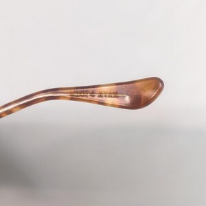 May include: Close-up of a brown and tan tortoiseshell eyeglass temple arm. The arm has a curved shape and a gold-colored metal hinge. The text on the arm is partially visible.