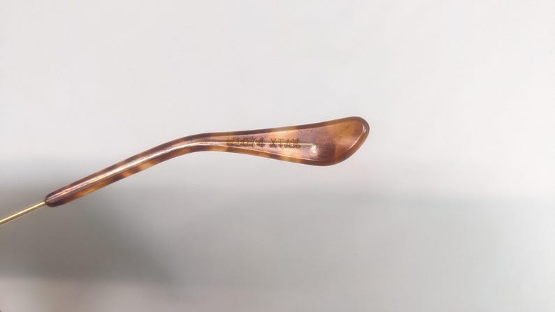 May include: Close-up of a brown and tan tortoiseshell eyeglass temple arm. The arm has a curved shape and a gold-colored metal hinge. The text on the arm is partially visible.