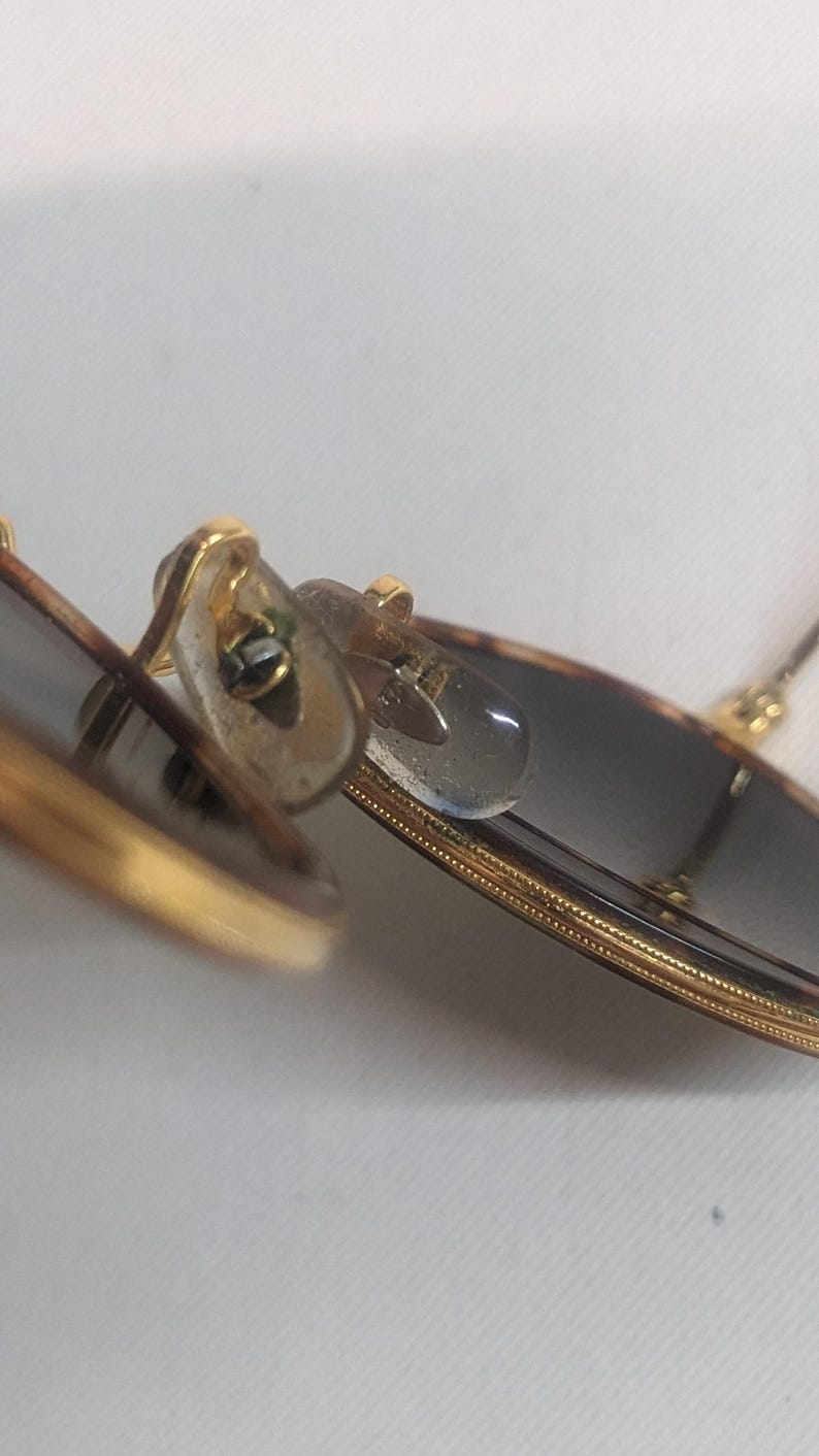 May include: Close-up of sunglasses with a gold frame and dark lenses. The nose pads are clear and the frame has a tortoiseshell pattern. The sunglasses are angled, showing the details of the frame and lenses.