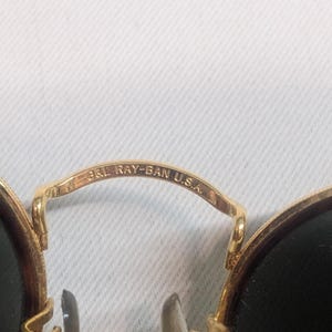 May include: Close-up of gold-rimmed sunglasses with dark lenses. The bridge of the glasses is engraved with "B&L RAY-BAN U.S.A." The sunglasses have a classic, round shape and are set against a white background.