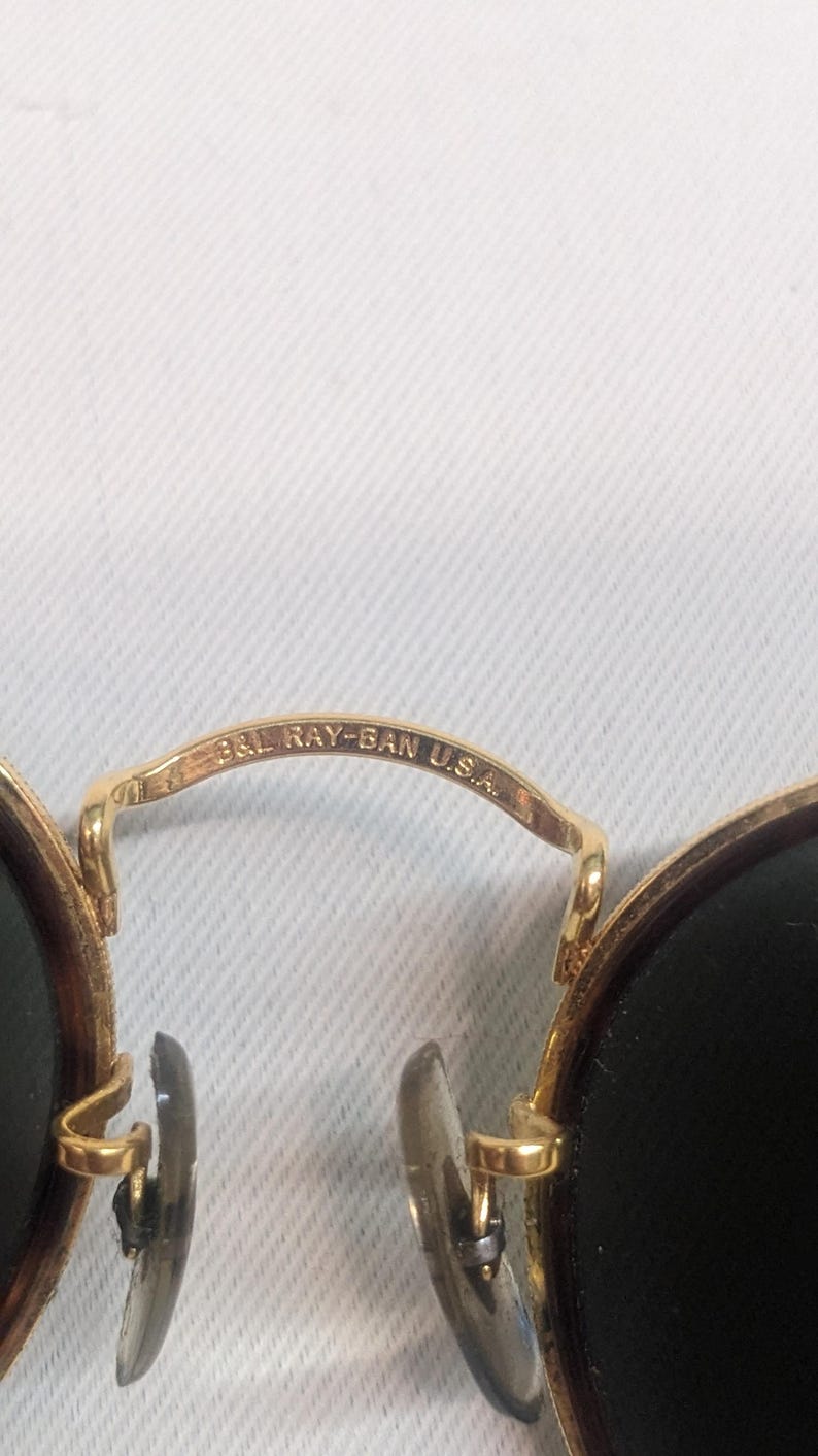 May include: Close-up of gold-rimmed sunglasses with dark lenses. The bridge of the glasses is engraved with "B&L RAY-BAN U.S.A." The sunglasses have a classic, round shape and are set against a white background.