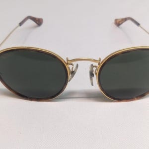 May include: Oval-shaped sunglasses with a gold-colored frame and dark green lenses. The rims have a tortoiseshell pattern. The temples are thin and gold, with tortoiseshell tips. The sunglasses are set against a white background.