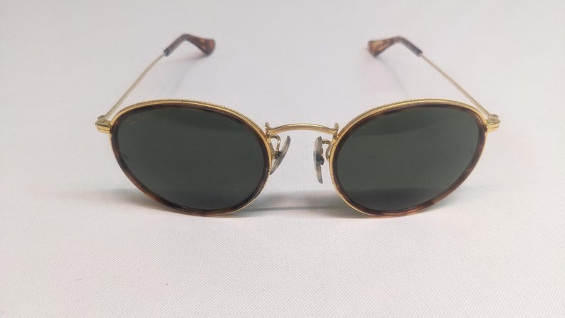 May include: Oval-shaped sunglasses with a gold-colored frame and dark green lenses. The rims have a tortoiseshell pattern. The temples are thin and gold, with tortoiseshell tips. The sunglasses are set against a white background.
