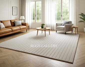 May include: A large, rectangular, light beige rug with a ribbed design dominates the room. A brown leather sofa with pillows, a gray armchair, and a round wooden coffee table are in the room. The text "RUGS GALLERY" is visible on the rug.