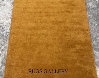 May include: Mustard-colored rectangular rug with a soft, plush texture. The rug has a solid color and the text "RUGS GALLERY" is printed in black near the bottom. The rug is likely made of a soft material.