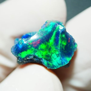 Puede incluir: Primer plano de una vibrante piedra preciosa de ópalo en bruto. La piedra muestra un llamativo juego de colores, con vívidos tonos verdes, azules y rojos arremolinándose por su superficie. El ópalo se sostiene en una mano enguantada.