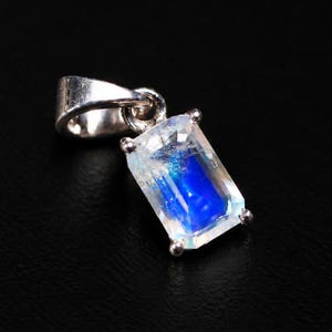 May include: A silver pendant featuring a rectangular, faceted moonstone with a prominent blue adularescence. The gemstone is secured by four metal prongs and attached to a polished silver bail, showcasing a clear, translucent appearance.