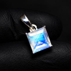 May include: A square-cut moonstone pendant set in a polished silver frame. The gemstone displays a translucent, milky white appearance with a distinct, glowing blue adularescence across its surface. The pendant includes a silver bail at the top.