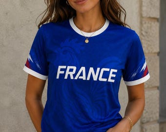 Puede incluir: Una camiseta azul con la palabra "FRANCE" en blanco. La camiseta tiene ribetes blancos en el cuello y las mangas, con detalles en rojo y blanco en las mangas. La camiseta se lleva con vaqueros azules.