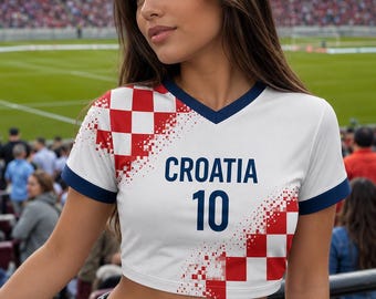 Puede incluir: Top corto blanco con diseño de bandera croata en las mangas y hombros. La camiseta tiene cuello en V y ribete azul marino, con la palabra "CROATIA" y el número "10" impresos en la parte delantera. Se usa con vaqueros azules.