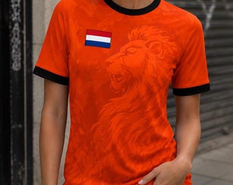 Puede incluir: Camiseta naranja con ribetes negros, con un gráfico de león y la bandera holandesa. La camiseta de manga corta tiene cuello redondo. La camiseta se usa con pantalones cortos de mezclilla.