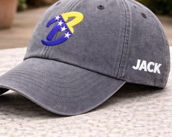 Puede incluir: Gorra de béisbol gris con visera curva. La gorra presenta una letra "B" estilizada en azul y amarillo con estrellas blancas. El nombre "JACK" está bordado en blanco en el lateral.