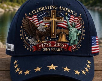 Puede incluir: Gorra de béisbol azul marino con estrellas doradas en la visera. La gorra presenta una bandera estadounidense, un águila, la Estatua de la Libertad y el texto "CELEBRATING AMERICA 1776-2026 250 YEARS".