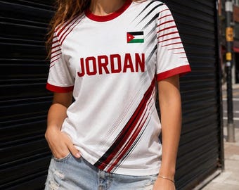 Puede incluir: Camiseta blanca con ribete rojo y la palabra "JORDAN" en letras rojas. La camiseta presenta una bandera jordana y rayas diagonales negras y rojas. La modelo lleva un pantalón corto vaquero.
