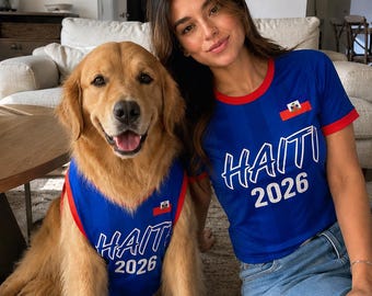 Puede incluir: Una mujer y un perro golden retriever llevan camisetas azules a juego con la palabra "HAITI" y el año "2026" impresos. Las camisetas tienen ribetes rojos y una pequeña bandera haitiana. La mujer lleva vaqueros azules.