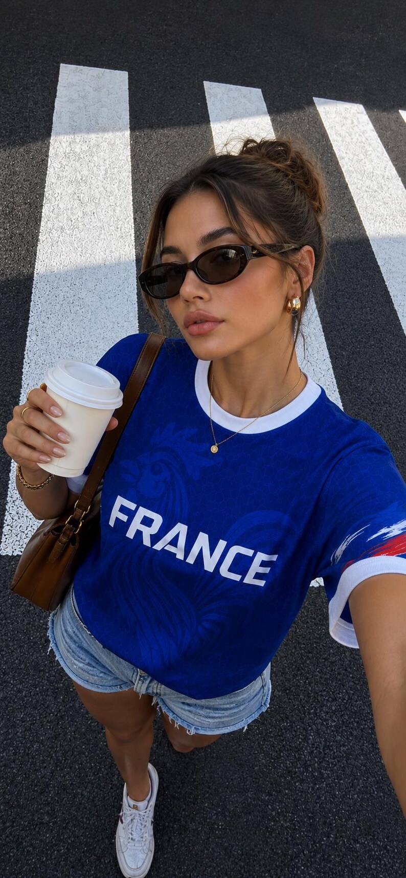 May include: A blue t-shirt with "FRANCE" printed on the front, white trim, and a brown shoulder bag. The person is wearing sunglasses and holding a white coffee cup. The background is a black and white crosswalk.